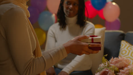 Frau-Gibt-Ihrer-Freundin-Zu-Hause-Ein-Geburtstagsgeschenk-Mit-Partyballons-Im-Hintergrund